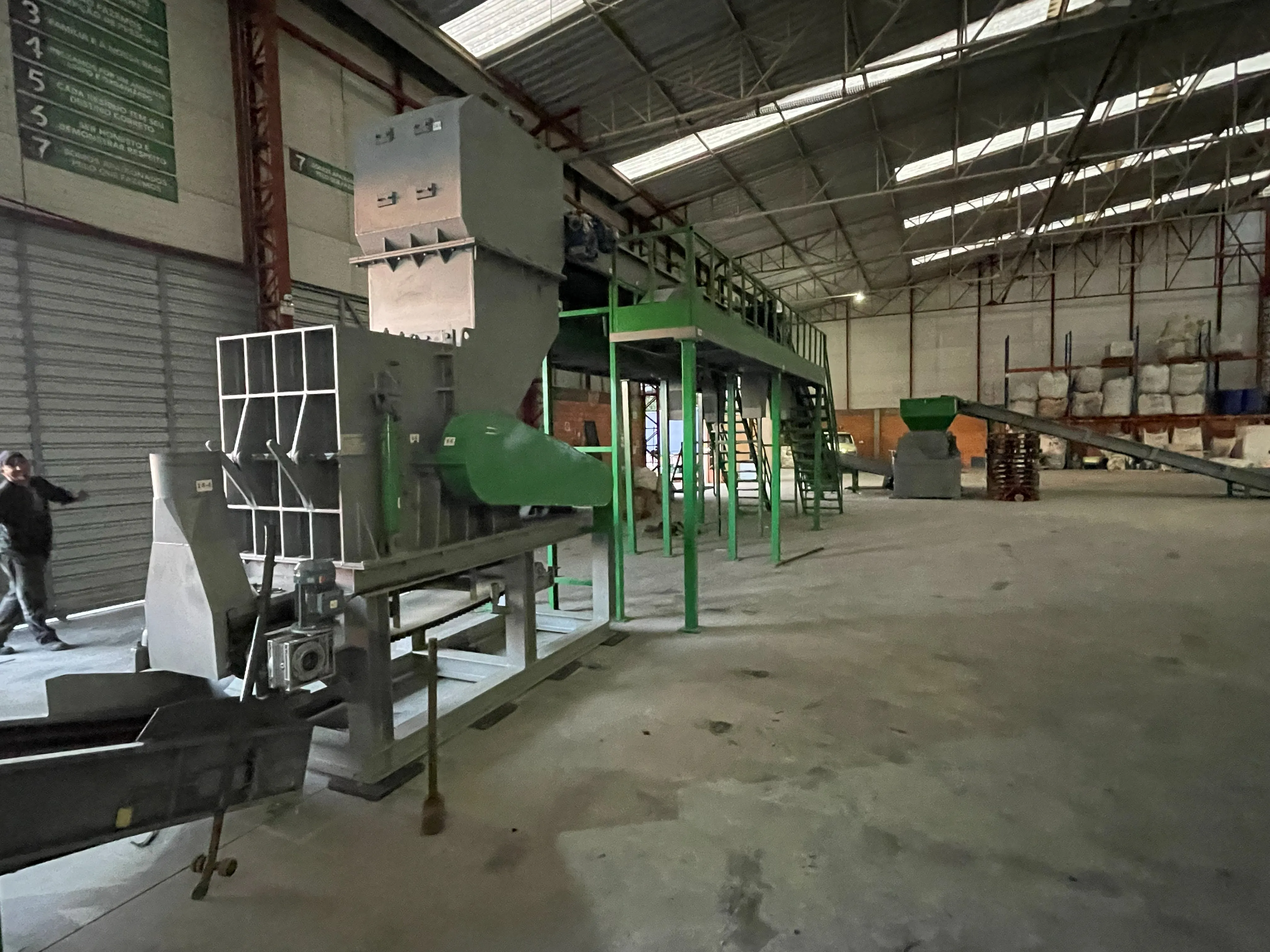 Linha de produção contínua instalada em galpão, com hopper cinza, plataforma e escada verdes.